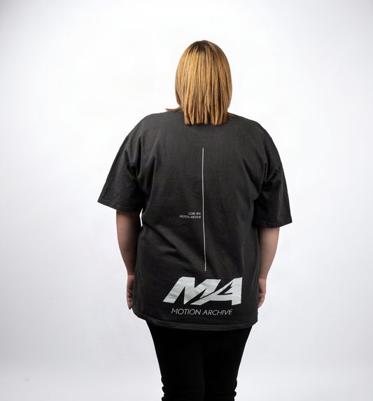 A person viewed from the back wearing an oversized black t-shirt with a white graphic print on the back. The print features a vertical line down the center, small text near the upper back, and a large stylized 'MA' logo with 'MOTION ARCHIVE' underneath.