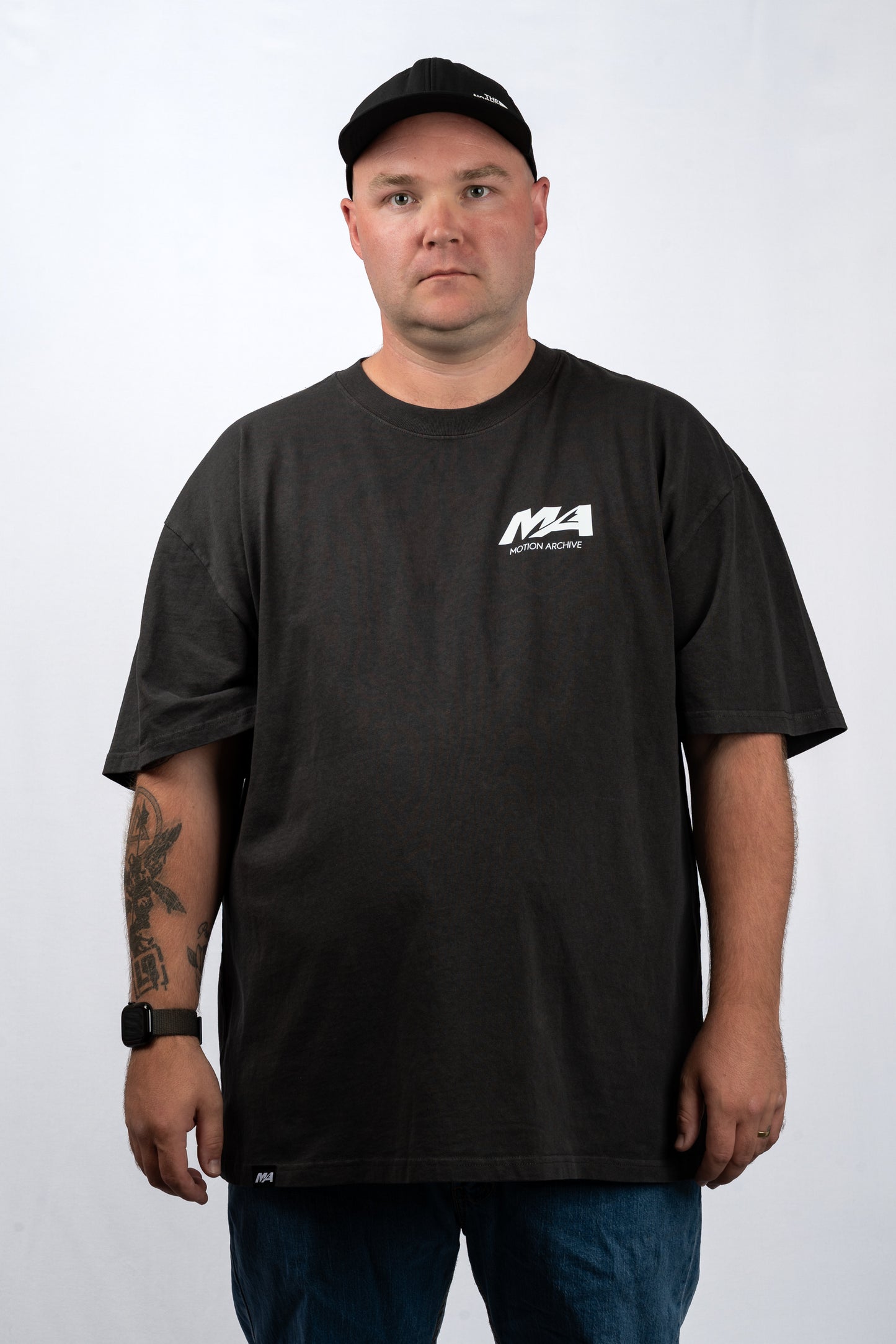A man wearing a black short-sleeve t-shirt with a white 'Moto Alpha' logo on the left chest, paired with a black baseball cap and blue jeans, standing against a light gray background.