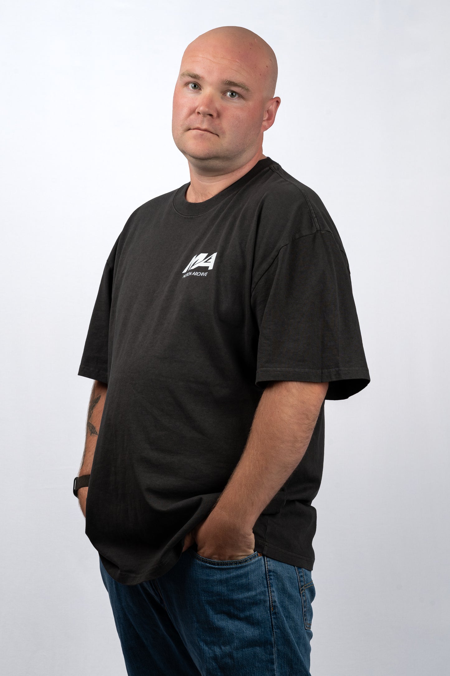 A bald man wearing a black crewneck t-shirt with a small white graphic logo on the left chest, paired with blue jeans, standing against a light gray studio background.