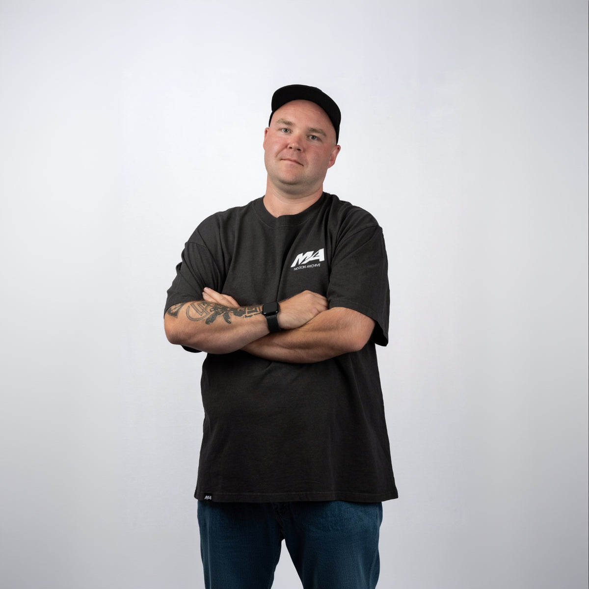 A man wearing a black crewneck t-shirt with a white 'M4' logo on the left chest, paired with blue jeans and a black baseball cap, standing with arms crossed against a light gray background.