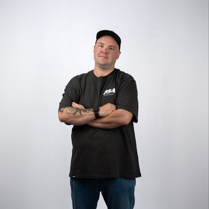 A man wearing a black crewneck t-shirt with a white 'M4' logo on the left chest, paired with blue jeans and a black baseball cap, standing with arms crossed against a light gray background.