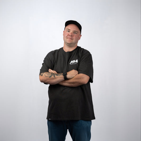 A man wearing a black crewneck t-shirt with a white 'M4' logo on the left chest, paired with blue jeans and a black baseball cap, standing with arms crossed against a light gray background.