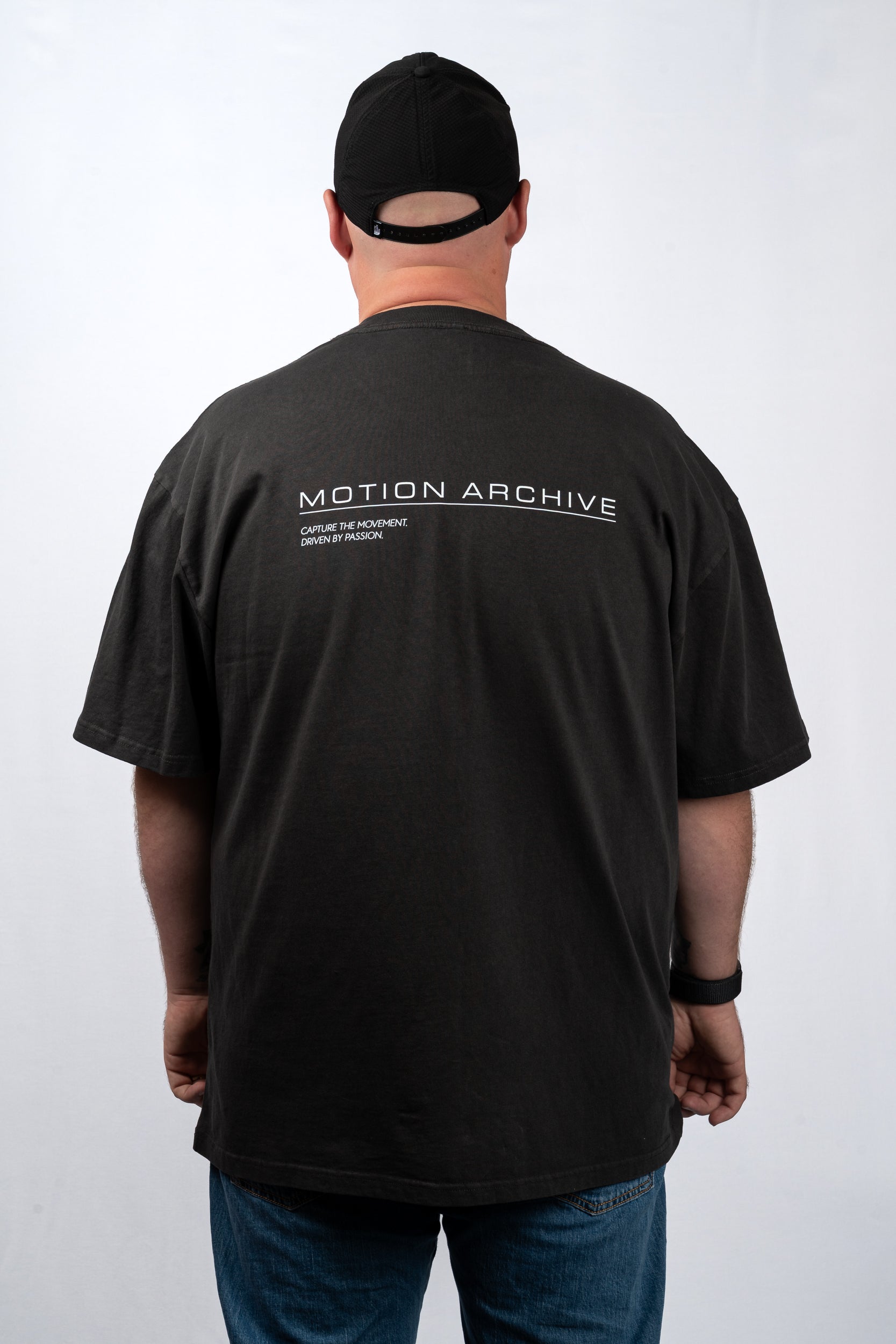A man viewed from behind wearing a black t-shirt with 'MOTION ARCHIVE' printed in white across the upper back, paired with a black baseball cap and blue jeans, against a light gray studio background.