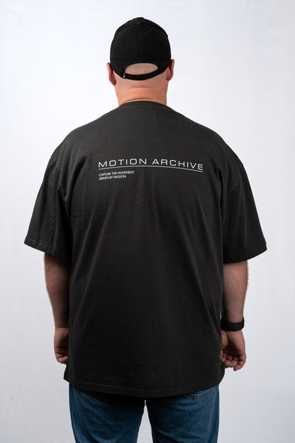 A man viewed from behind wearing a black t-shirt with 'MOTION ARCHIVE' printed in white across the upper back, paired with a black baseball cap and blue jeans, against a light gray studio background.