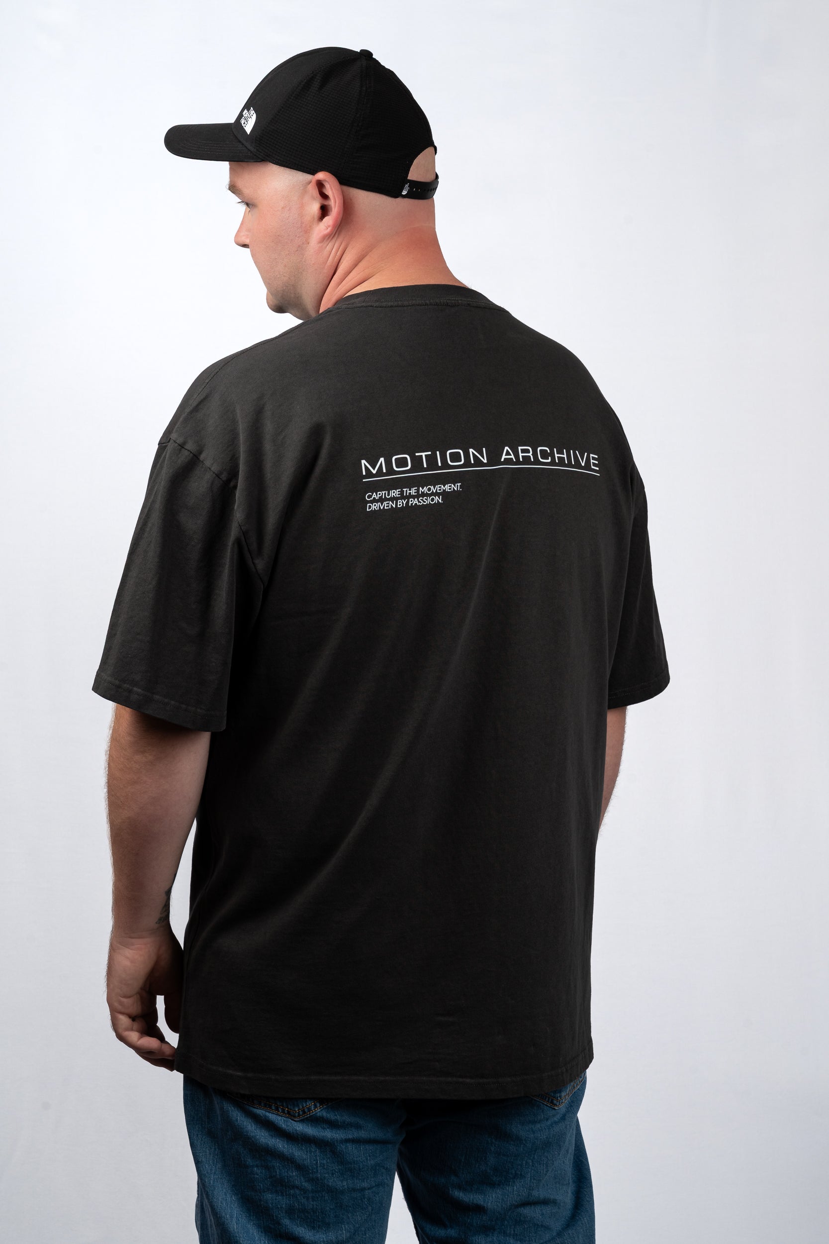 A man wearing a black t-shirt from the back, featuring 'MOTION ARCHIVE' printed in white across the upper back, paired with a black baseball cap and blue jeans against a light gray background.