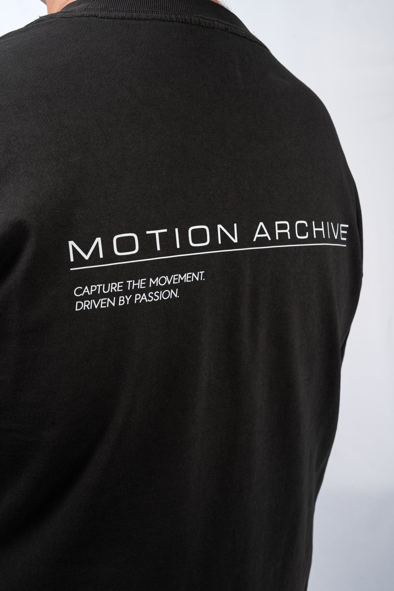 Close-up of the back of a black short-sleeve t-shirt featuring white printed text that reads 'MOTION ARCHIVE' and 'CAPTURE THE MOVEMENT. DRIVEN BY PASSION.