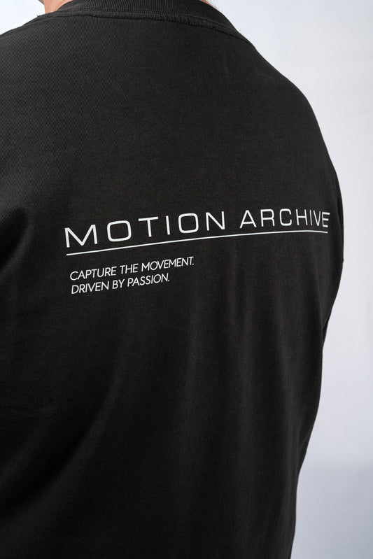 Close-up of the back of a black short-sleeve t-shirt featuring white printed text that reads 'MOTION ARCHIVE' and 'CAPTURE THE MOVEMENT. DRIVEN BY PASSION.