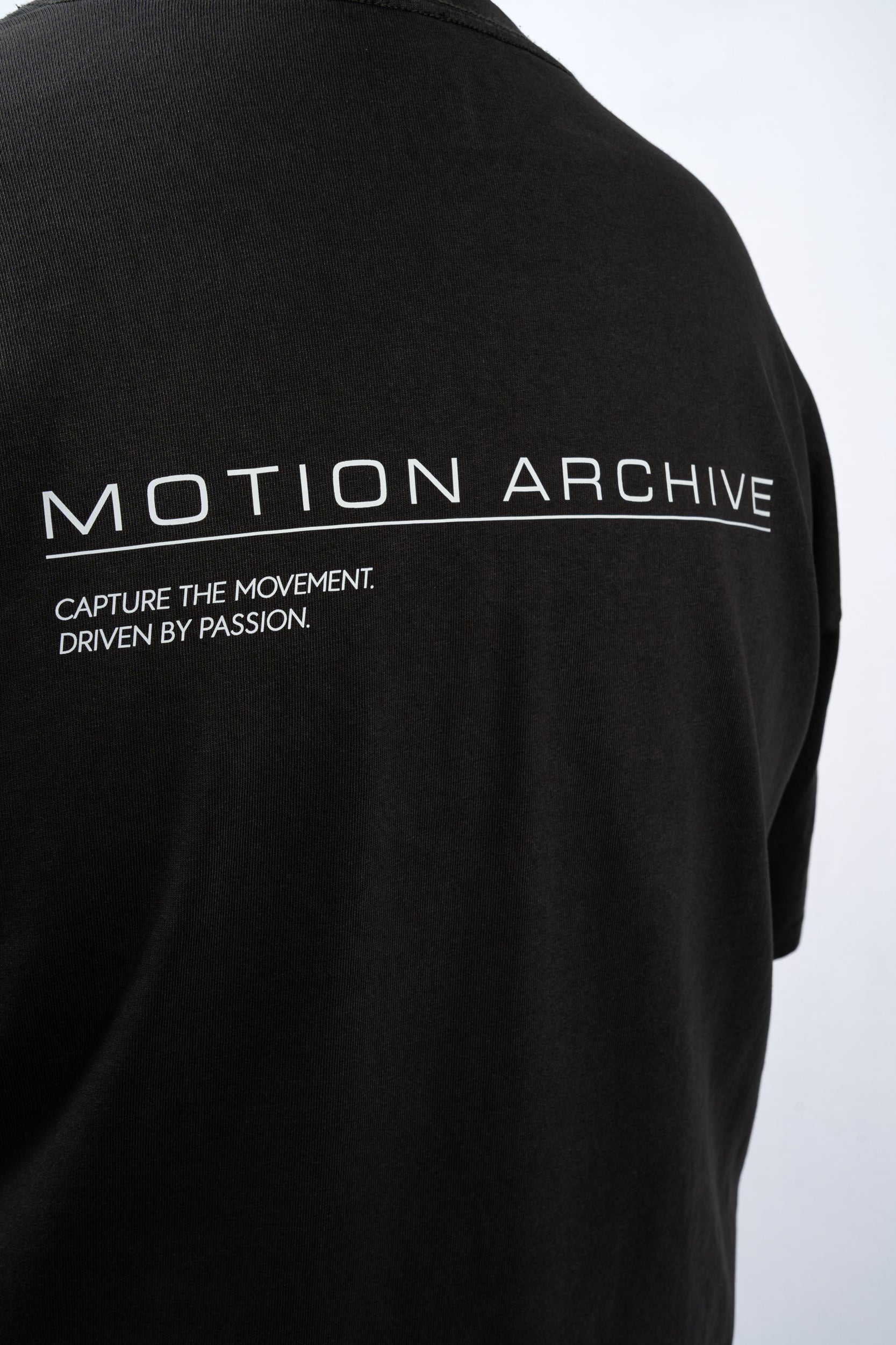 Close-up of the back of a black short-sleeve t-shirt featuring white printed text that reads 'MOTION ARCHIVE' and 'CAPTURE THE MOVEMENT. DRIVEN BY PASSION.