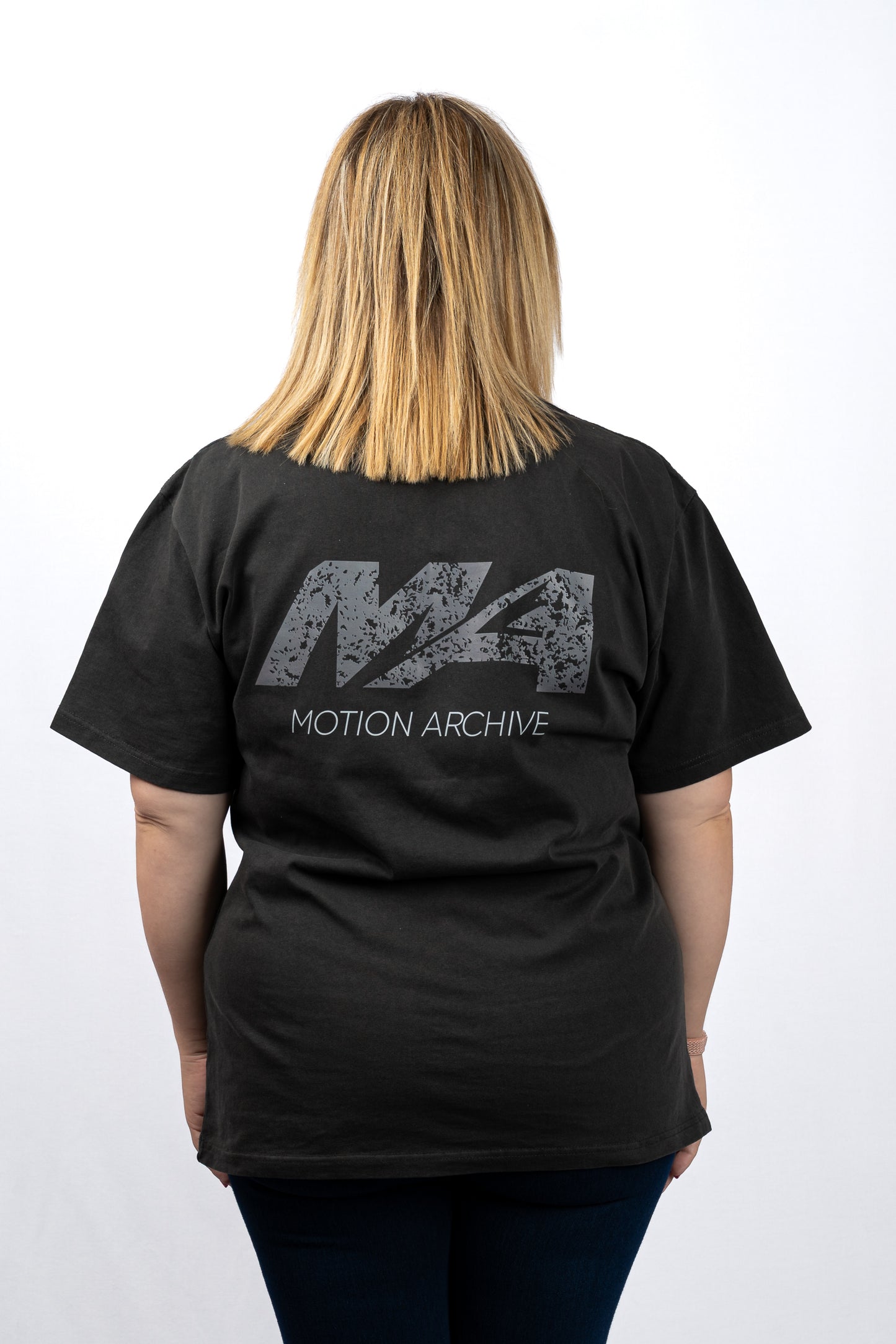 A woman viewed from behind wearing a black t-shirt with a distressed gray 'M4' logo and 'MOTION ARCHIVE' text printed on the back, set against a plain white background.