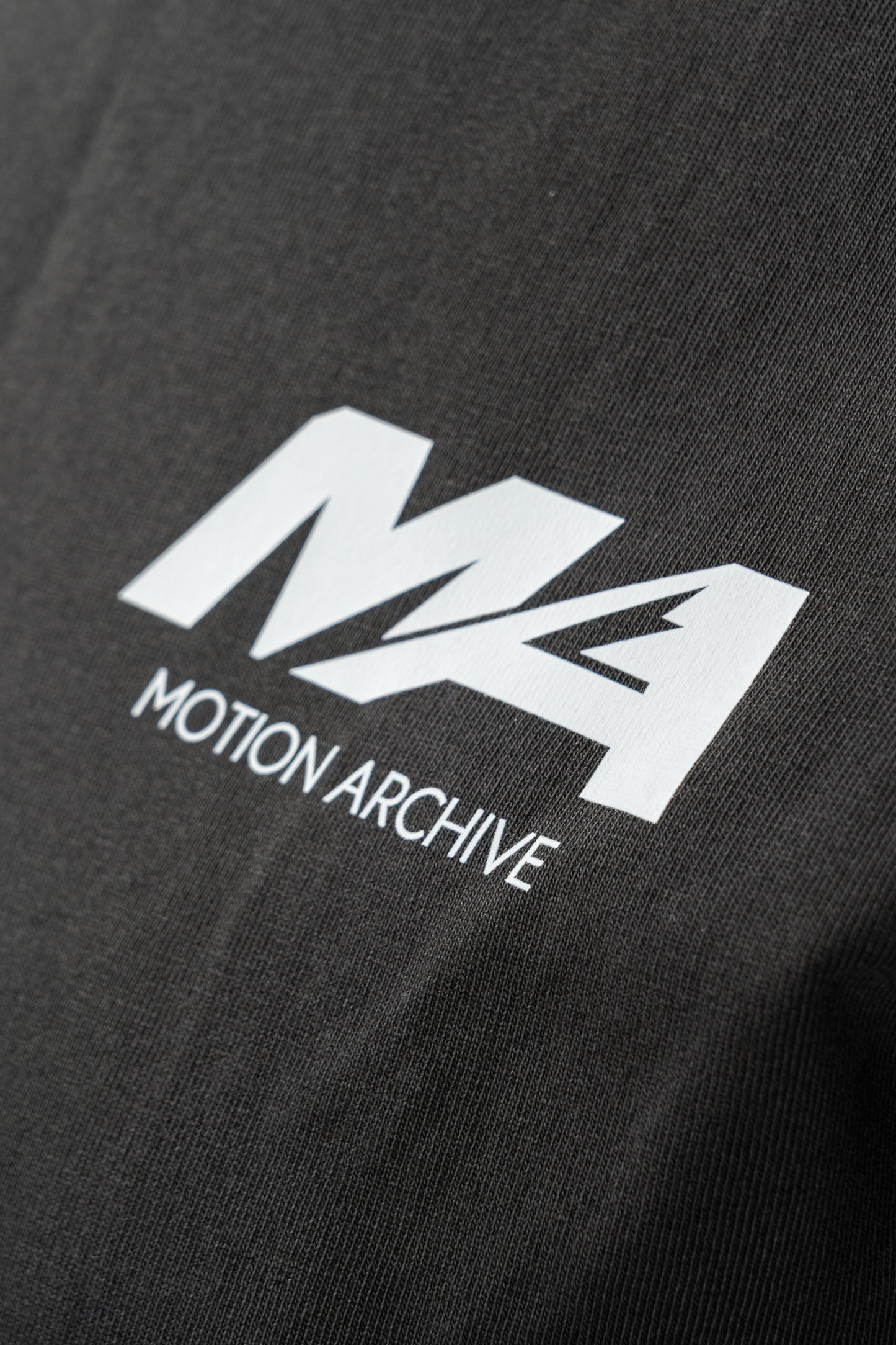 Close-up of a black t-shirt featuring a white printed logo with the text 'M4 MOTION ARCHIVE'.