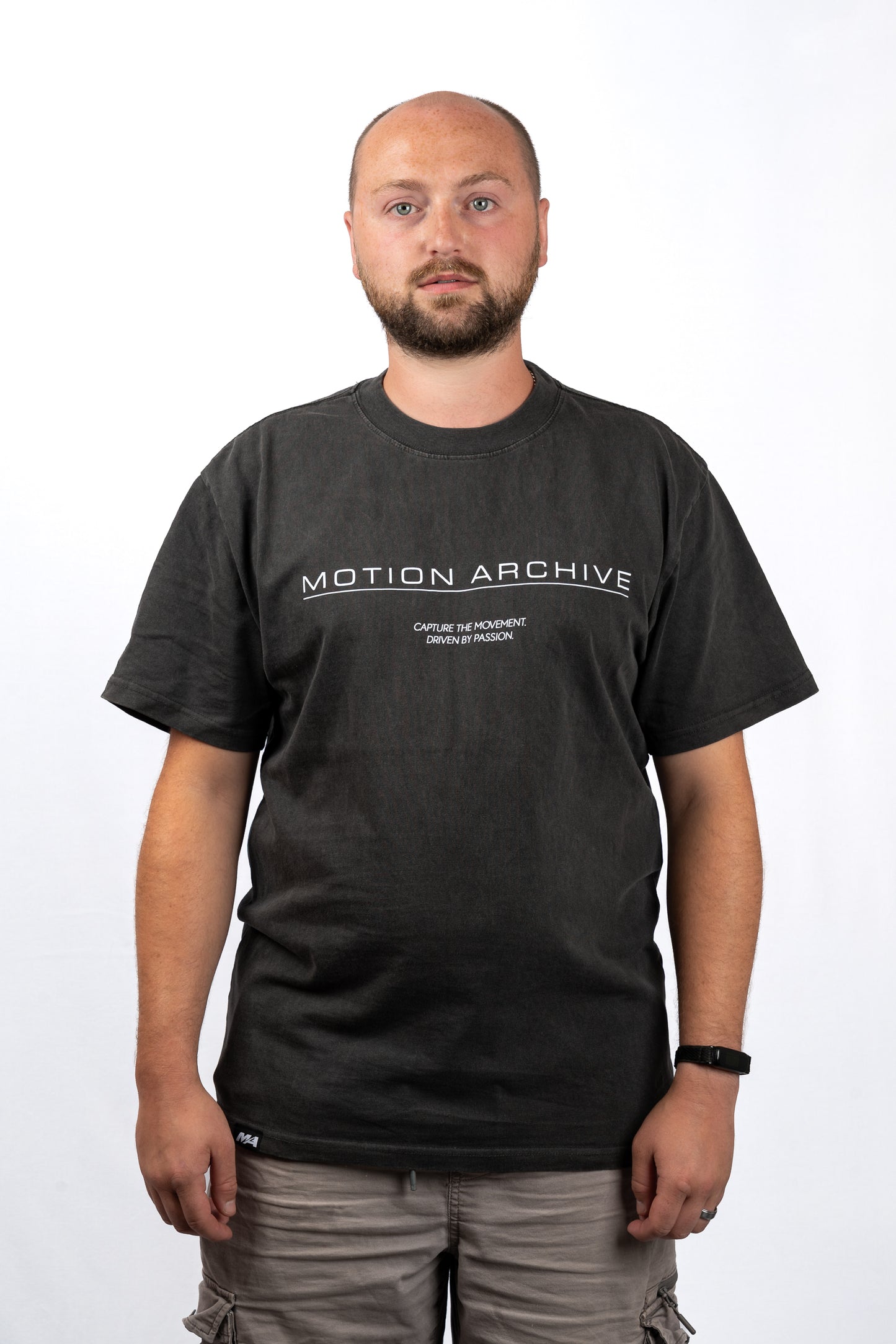 A man wearing a dark charcoal gray t-shirt with 'MOTION ARCHIVE' printed in white across the chest, paired with gray cargo pants, photographed against a plain white background.