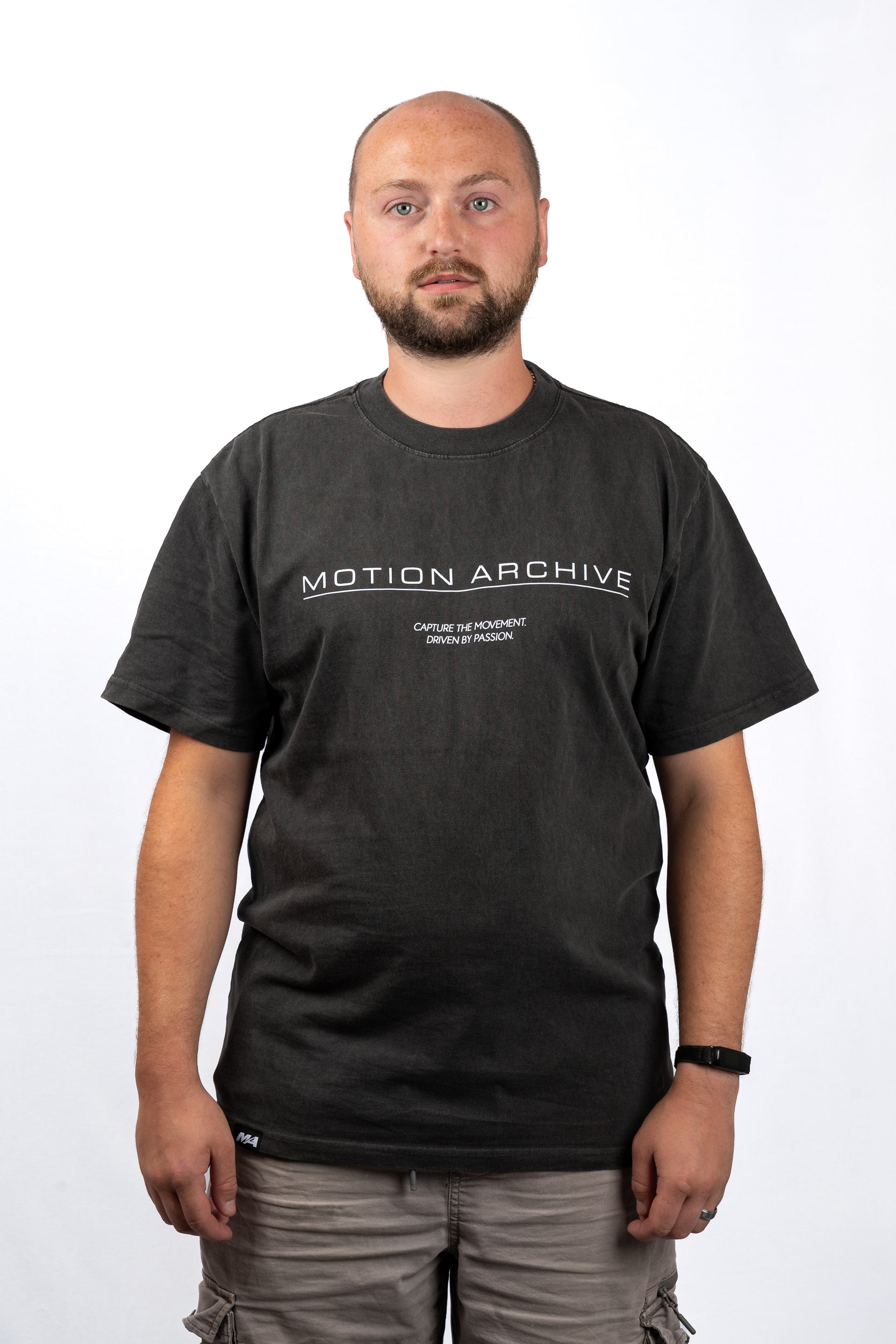 A man wearing a dark charcoal gray t-shirt with 'MOTION ARCHIVE' printed in white across the chest, paired with gray cargo pants, photographed against a plain white background.