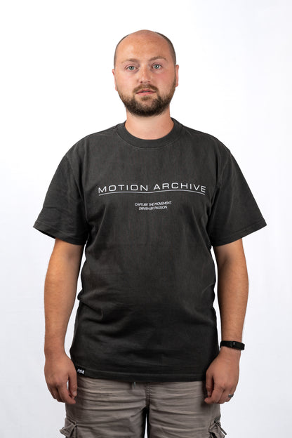 A man wearing a dark charcoal gray t-shirt with 'MOTION ARCHIVE' printed in white across the chest, paired with gray cargo pants, photographed against a plain white background.