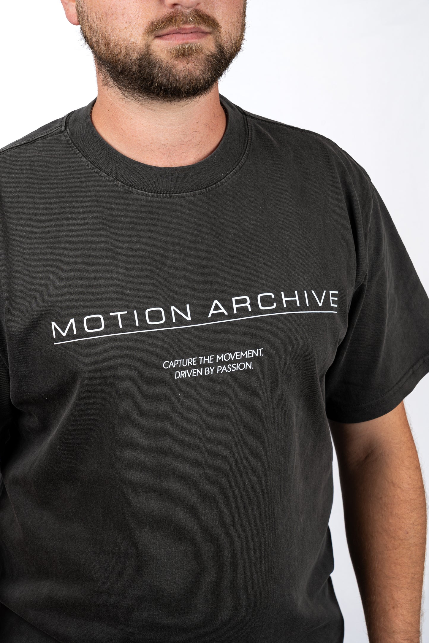 A man wearing a dark charcoal gray t-shirt with 'MOTION ARCHIVE' printed in white across the chest, shown from the chin down against a plain white background.