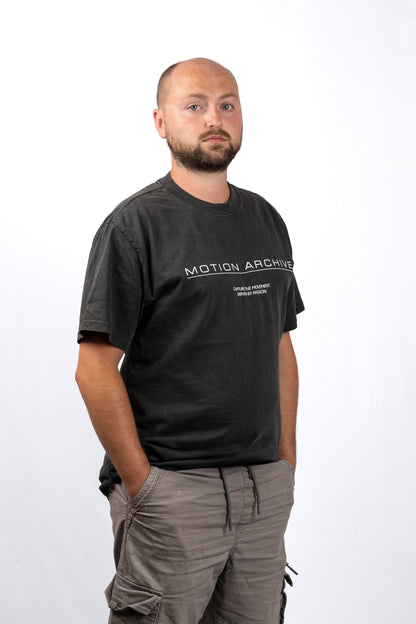 A man wearing a faded black t-shirt with 'MOTION ARCHIVE' printed in white across the chest, paired with gray cargo pants, standing against a plain white background.
