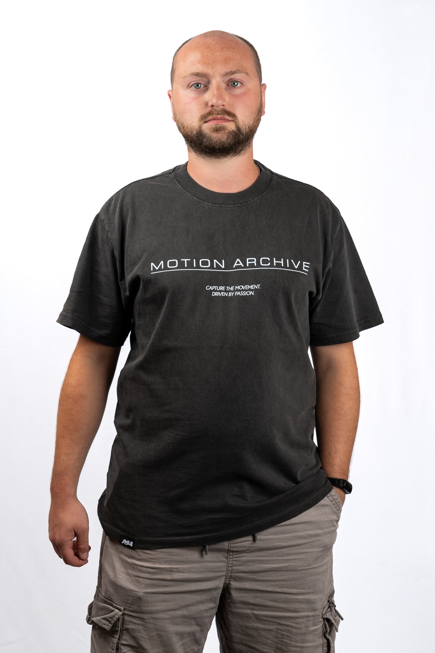 A man wearing a dark charcoal gray t-shirt with 'MOTION ARCHIVE' printed in white across the chest, paired with gray cargo pants, photographed against a plain white background.