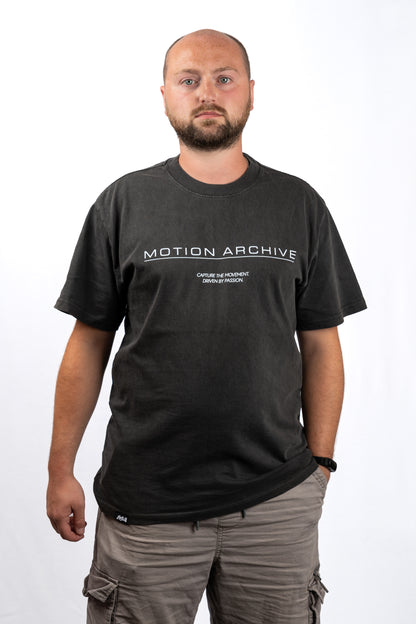 A man wearing a dark charcoal gray t-shirt with 'MOTION ARCHIVE' printed in white across the chest, paired with gray cargo pants, photographed against a plain white background.