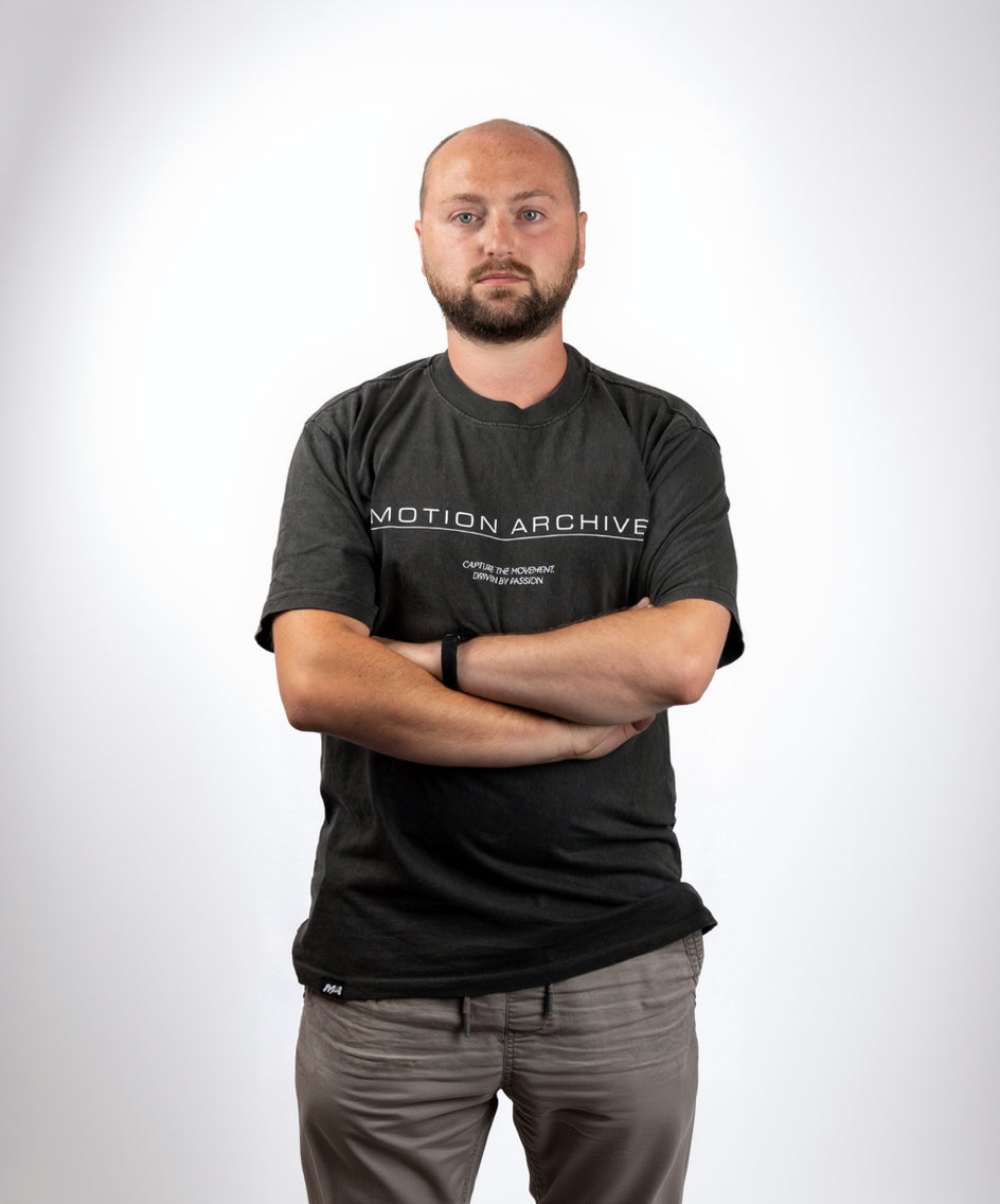 A man with a shaved head and beard wearing a dark gray t-shirt with 'MOTION ARCHIVE' printed in white across the chest, standing with arms crossed against a plain white background.
