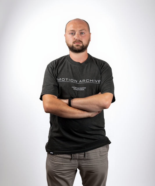 A man with a shaved head and beard wearing a dark gray t-shirt with 'MOTION ARCHIVE' printed in white across the chest, standing with arms crossed against a plain white background.