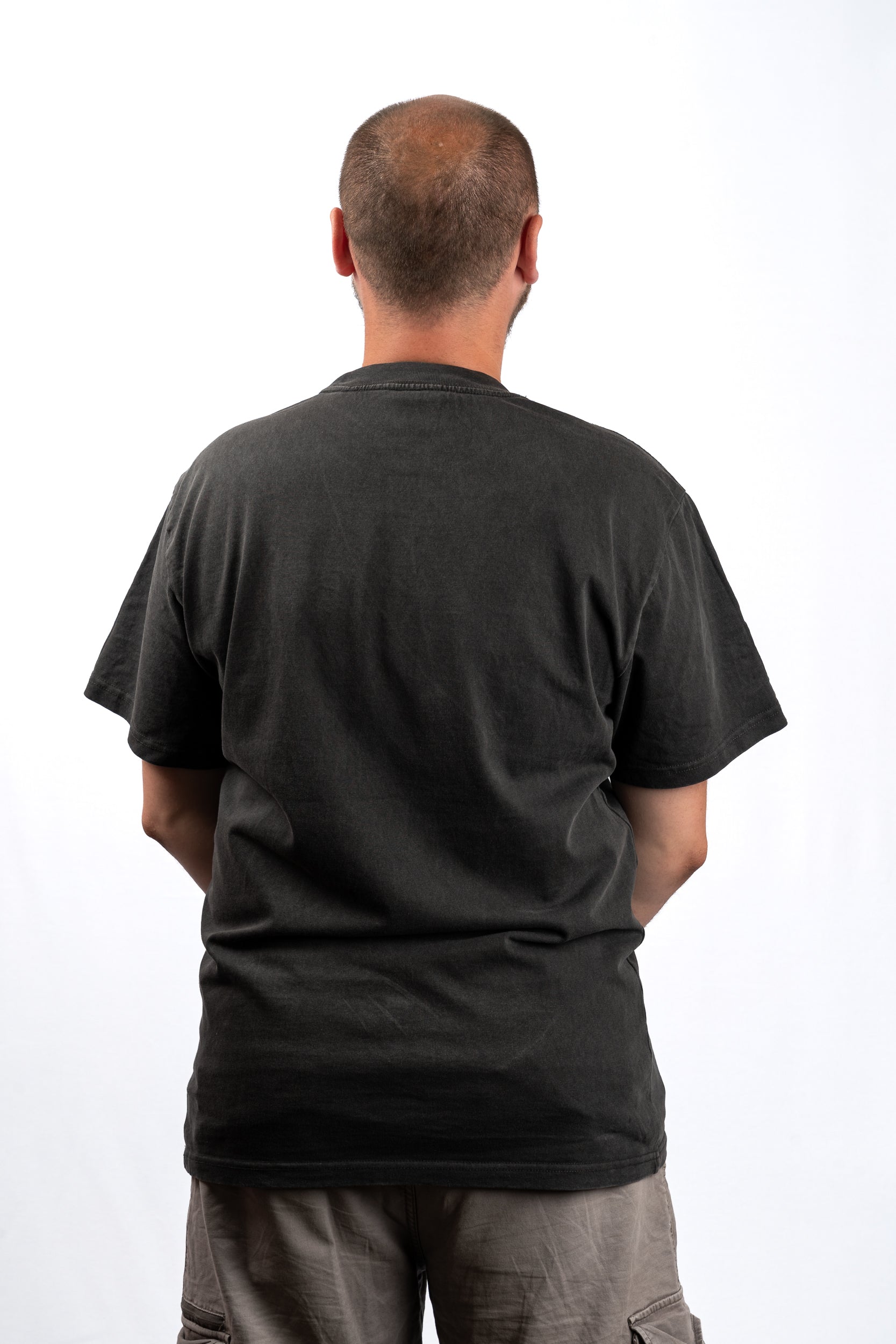 A man viewed from behind wearing a plain black short-sleeve t-shirt and gray cargo pants against a white studio background.