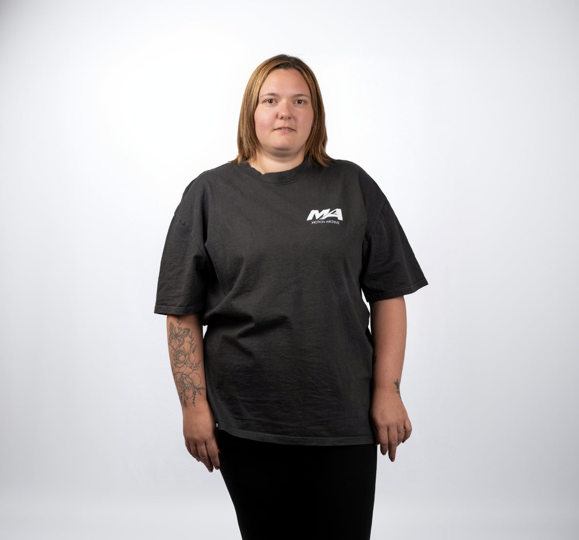 A woman with shoulder-length blonde hair stands against a light gray background wearing a dark charcoal gray t-shirt with a small white 'MA' logo on the left chest, paired with black pants.