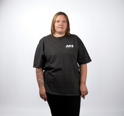 A woman with shoulder-length blonde hair stands against a light gray background wearing a dark charcoal gray t-shirt with a small white 'MA' logo on the left chest, paired with black pants.