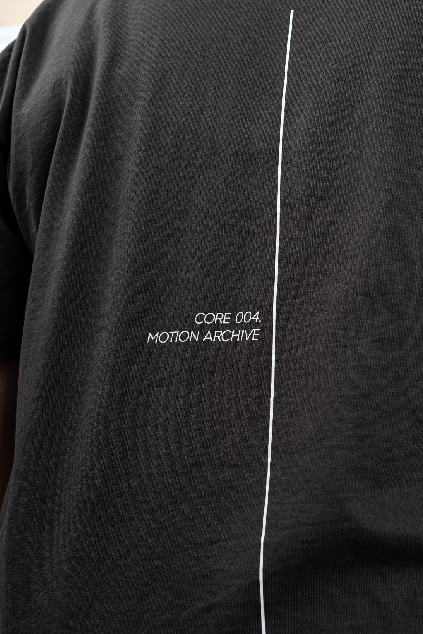 Close-up of a black short-sleeve t-shirt featuring a vertical white line down the center and the white text 'CORE 004. MOTION ARCHIVE' printed on the chest.