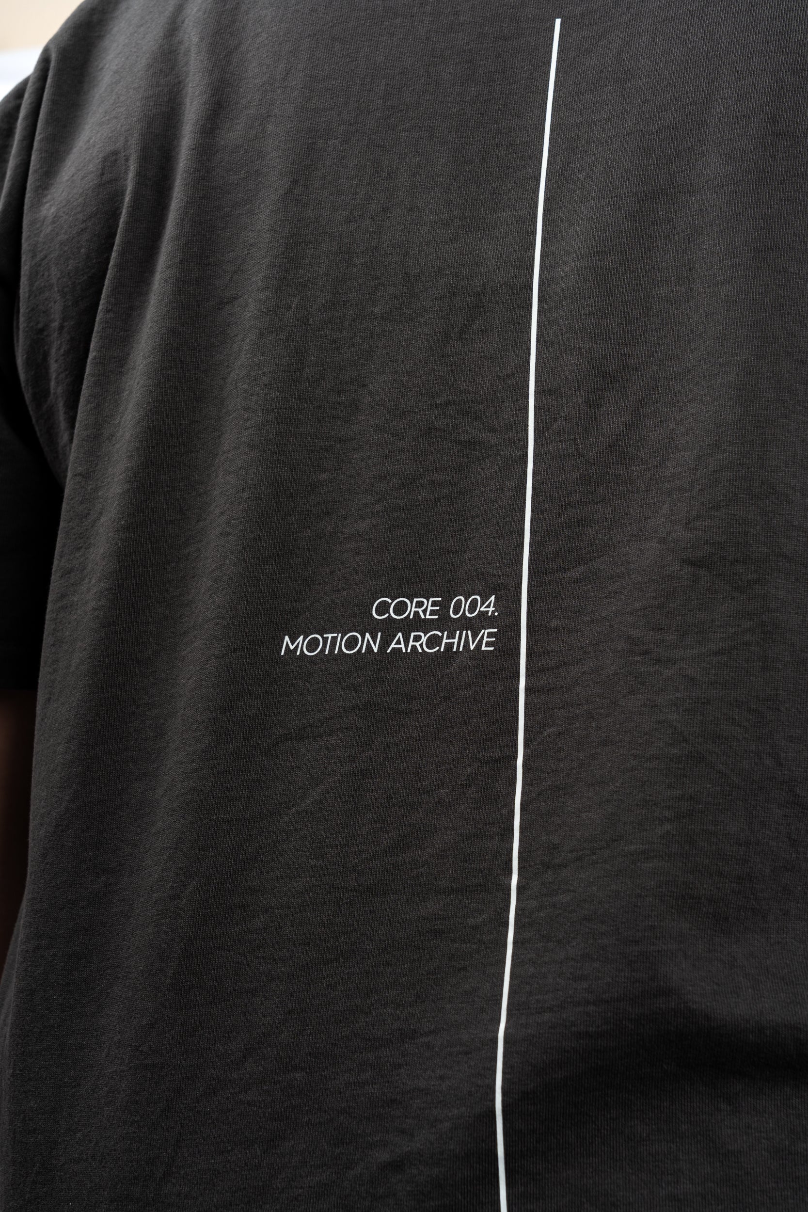 Close-up of a black short-sleeve t-shirt featuring a vertical white line down the center and the white text 'CORE 004. MOTION ARCHIVE' printed on the chest.