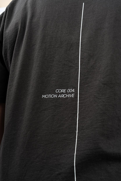Close-up of a black short-sleeve t-shirt featuring a vertical white line down the center and the white text 'CORE 004. MOTION ARCHIVE' printed on the chest.