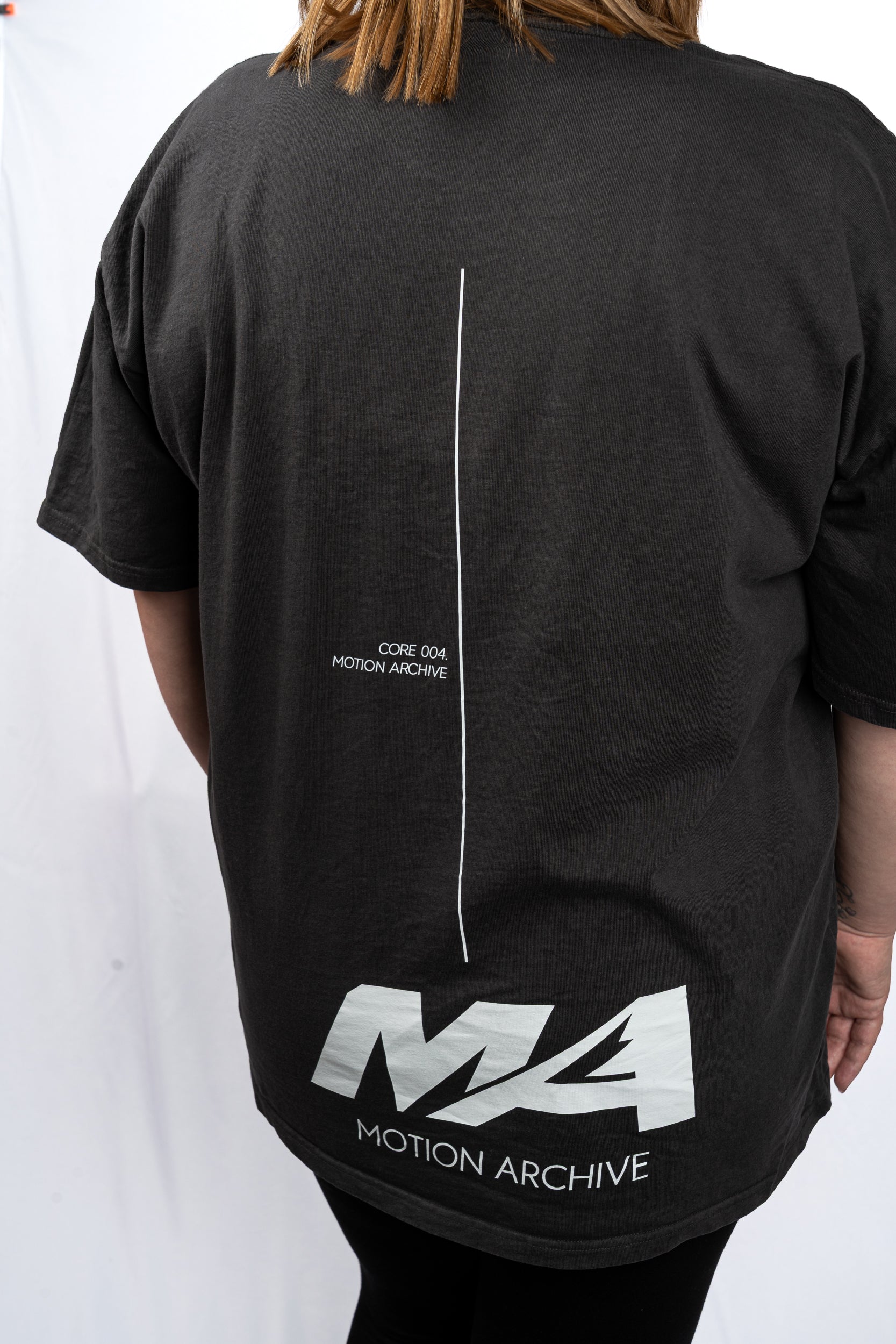 A person shown from the back wearing an oversized black t-shirt with a white vertical line graphic and 'MA MOTION ARCHIVE' branding printed on the back.
