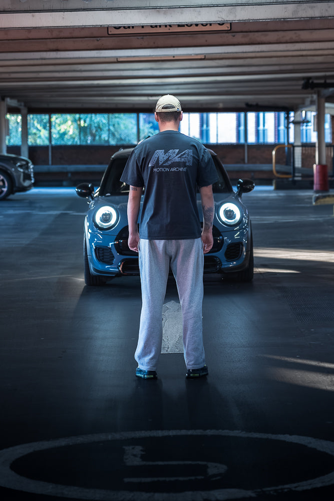 A man stands with his back to the camera in a parking garage, facing a blue car with its headlights on. He is wearing a dark gray t-shirt with a white 'M4 Motorsport' logo on the back, white sweatpants, and a beige baseball cap.